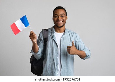 Happy black man showing thumb up and flag of France, carrying backpack on shoulder, grey studio background, copy space. Smiling african american guy student with French flag, education in France