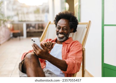 Happy black man sitting outdoors on a deck chair, using his mobile phone for a video call. Mature man connecting with his friends and family in a casual and relaxed moment on his patio. - Powered by Shutterstock