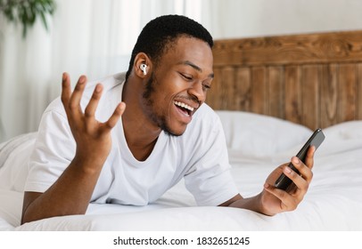 Happy Black Man With Smartphone Making Video Call Online Using Earbuds Earphones Lying In Bed At Home. Distant Mobile Communication, Video Conference Application Concept