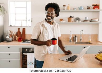 Happy black man standing in his kitchen at home, drinking a cup of coando café, traditional Brazilian coffee. Mature man enjoying his morning with a hot beverage and a laptop for internet browsing. - Powered by Shutterstock