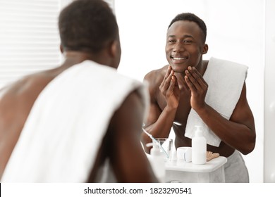 Happy Black Man Touching Smooth Face Skin After Shaving Standing Near Mirror In Bathroom At Home. Male Facial Skincare, Morning Beauty Routine And Self-Care Cosmetics Concept