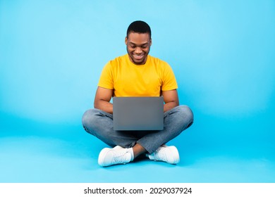 Happy Black Man Using Laptop Working Online Sitting Over Blue Background In Studio. Successful African American Male Freelancer Or Blogger Guy Browsing Internet On Computer