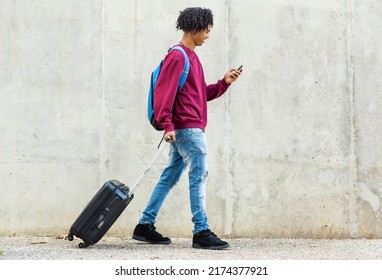 Happy Black Man Using Mobile Phone While Pulling Rolling Suitcase Outdoors In City