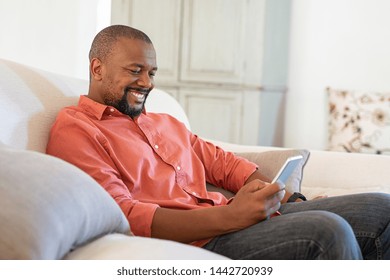 Happy black man using smart phone while relaxing at home. Smiling mature man at home sitting on couch reading phone message. African american guy download app for his cellphone.