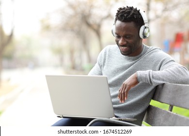 Happy Black Man Watching Media On Laptop Wearing Headphones Sitting On A Bench In A Park