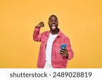 Happy Black man winner holding smartphone celebrating win isolated on yellow. Excited African young gen z guy holding cellphone. Amazed user sport betting game app winner using mobile phone.