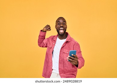 Happy Black man winner holding smartphone celebrating win isolated on yellow. Excited African young gen z guy holding cellphone. Amazed user sport betting game app winner using mobile phone. - Powered by Shutterstock