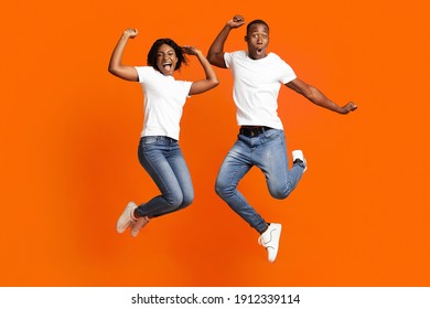 Happy Black Man And Woman Celebrating Success, Full Length Photo, Copy Space. Cheerful African American Young Couple Clenching Fists And Screaming, Jumping Up Over Orange Studio Background