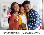 Happy black military man wrapped in american flag posing with wife and little daughter at home after returning from army, cheerful family celebrating reunion, embracing and smiling at camera