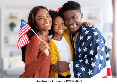 Happy black military man wrapped in american flag posing with wife and little daughter at home after returning from army, cheerful family celebrating reunion, embracing and smiling at camera - Powered by Shutterstock