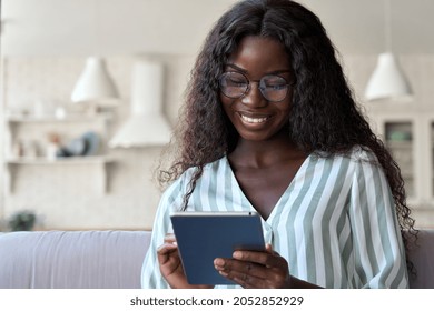 Happy black mixed raced African black young woman in glasses with perfect smile sitting on sofa holding using tablet device reading e book, surfing social media dating apps in apartment.