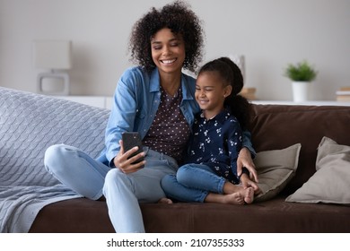 Happy Black Mom And Cheerful Kid Taking Selfie On Smartphone, Making Video Call, Relaxing On Home Couch, Enjoying Leisure Together. Young Mother And Little Daughter Using Mobile Phone, Laughing