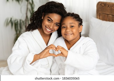 Happy Black Mom And Daughter In Bathrobes Making Heart Sign Form Hands, Connecting Their Arms, Having Fun And Bonding At Home, Relaxing On Bed Together After Bath, Smiling At Camera, Closeup