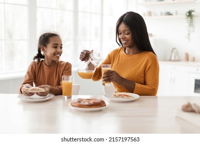 Happy Black Mom And Her Daughter Drinking Fresh Orange Juice, Pouring It Filling Glasses During Lunch Sitting At Table In Kitchen At Home. Family Nutrition Concept