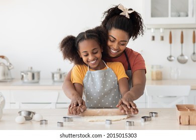 Happy Black Mom Showing Her Teen Kid Hot To Bake Cookies, Cheerful African American Mother And Daughter Rolling Dough For Homemade Pastry And Smiling, Wearing Aprons, Kitchen Interior