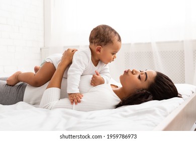 Happy Black Mommy Playing With Toddler Baby Lying On Her Stomach Posing In Bedroom At Home, Side View. Loving Mother Talking To Little Son Spending Time With Child During Maternity Leave. Motherhood