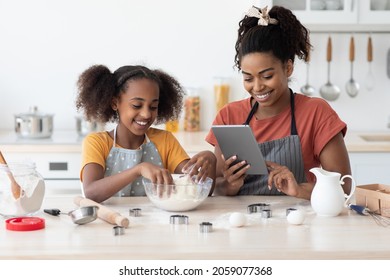 Happy Black Mother And Daughter Baking Together At Home, Reading Culinary Blog On Internet, Using Digital Tablet, African American Mom And Teen Kid Making Dough For Cookies Or Cake