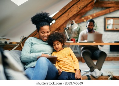 Happy black mother and daughter relaxing on the sofa and surfing the net on touchpad at home. Father is working on laptop in the background.