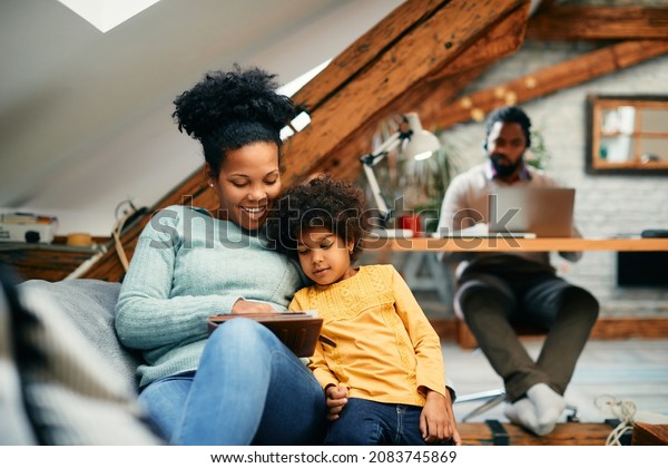 Happy black mother and daughter relaxing on\
the sofa and surfing the net on touchpad at home. Father is working\
on laptop in the\
background.