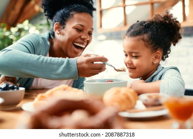 Happy black mother having fun and feeding her daughter while having breakfast at dining table. - Shutterstock ID 2087887318