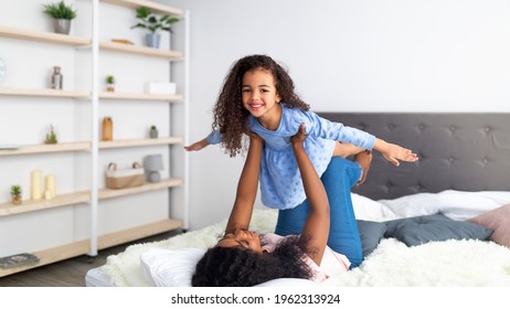 Happy Black Mother And Her Cute Daughter Playing Airplane Game On Bed At Home, Panorama. Lovely Little African American Girl Having Fun With Her Mom, Being Silly. Family Bonding Times