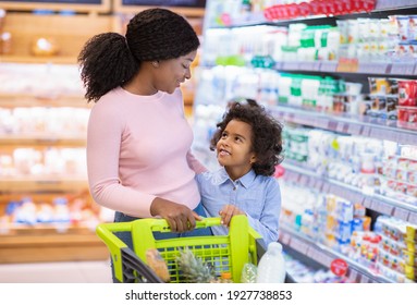 Happy black mother with her daughter shopping for groceries, selecting dairy products at mall. Young African American family with trolley buying food at huge hypermarket