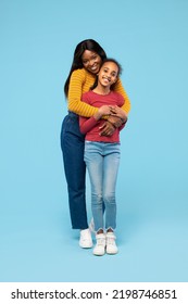 Happy Black Mother Hugging Her Lovely Daughter Standing In Studio Over Blue Background, Full Length Shot. Young Mom And Her Kid Girl Bonding And Smiling To Camera