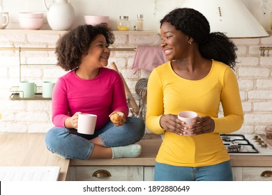 Happy Black Mother And Little Daughter Talking And Eating Snacks In Kitchen, Enjoying Homemade Pastry And Hot Cocoa, Cute African American Girl And Her Mom Having Fun Together At Home