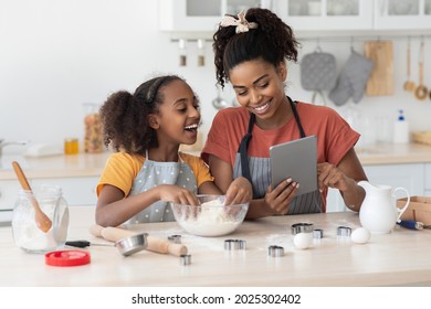 Happy black mummy and daughter cooking at home together, wearing aprons, standing by kitchen table with dough, flour, rolling pin and cookies figurines, using tablet, reading culinary blog