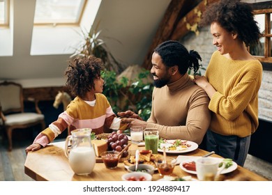 Happy black parents and their small daughter communicating during a meal in dining room. 