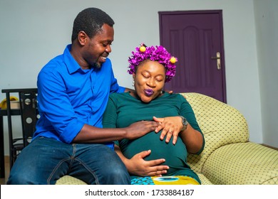 A happy black pregnant couple expecting delivery in covid-19 pandemic season - Happy African couple feeling fetus kick in mother's abdomen - maternity shoot indoors during quarantine or lock down 