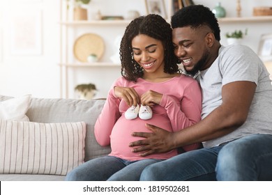 Happy black pregnant couple placing baby shoes on woman big belly while resting together on couch at home. Joyful african american young family waiting for baby, playing with baby boots, copy space