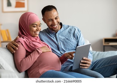 Happy Black Pregnant Muslim Couple Relaxing With Digital Tablet At Home, Smiling Islamic Spouses Shopping Online, Purchasing Clothes For Baby In Internet While Resting On Couch In Living Room