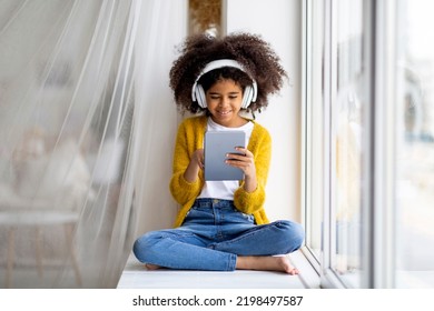 Happy Black Preteen Girl Enjoying Brand New Digital Tablet, Child Sitting On Windowsill At Home, Holding Pad, Using Wireless Headphones, Checking Newest Mobile Applications For Kids, Copy Space