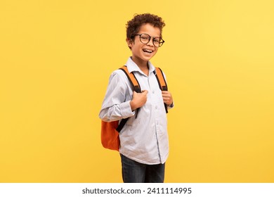 Happy black schoolboy with curly hair, wearing glasses and smiling at camera, carrying an orange backpack on sunny yellow background - Powered by Shutterstock