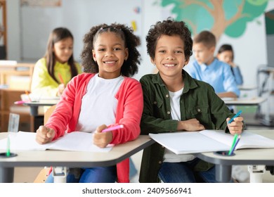 Happy black schoolboy and schoolgirl looking at camera and smiling, sitting at desks with other children on background - Powered by Shutterstock