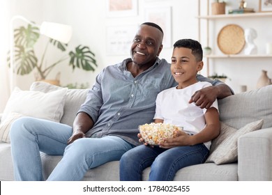 Happy Black Senior Man Spending Time With His Grandson At Home, Watching Tv And Eating Popcorn Together