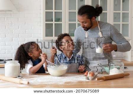 Happy Black sibling kids and young dad wearing aprons, cooking bakery food for family dinner together. Father teaching kids to bake, mixing dough in bowl for pastry, use flour, milk, eggs, whiskers