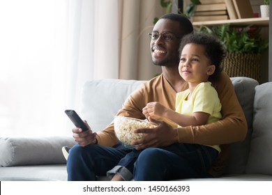 Happy Black Single Father And Cute Little Son Sit On Couch Relax Watching TV Eating Popcorn Together, Smiling African American Dad And Preschooler Child Enjoy Family Weekend With Snack And Cartoon