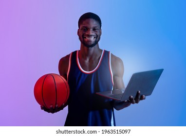 Happy Black Sports Fan Holding Laptop Computer And Basketball In Neon Light. Handsome African American Guy With Pc And Ball Cheering For His Favorite Team Online, Smiling At Camera