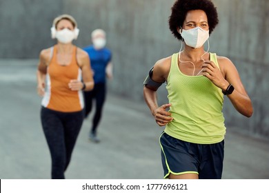 Happy black sportswoman wearing protective face mask while running outdoors during COVID-19 epidemic. There are people in the background. 