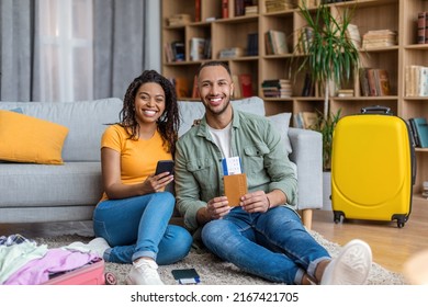 Happy Black Spouses Booking Tour Online, Using Cellphone, Man Holding Passport With Tickets, Smiling Together To Camera. Couple Contacting Tourist Agency On Web