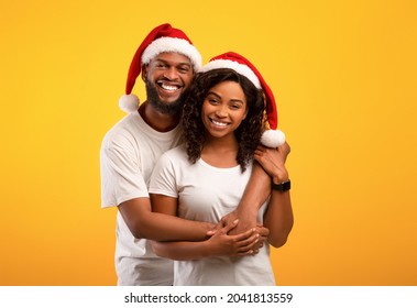Happy black spouses in Santa hats hugging on yellow studio background. Excited african american guy embracing his pretty wife from behind, celebrating New Year or Christmas together