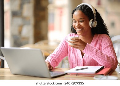 Happy black student e-learning with a laptop and headphone drinking coffee in a bar - Powered by Shutterstock