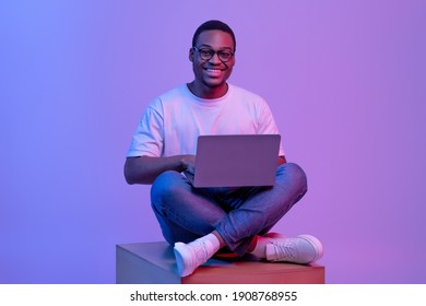 Happy Black Student Guy In Eyeglasses Sitting With Laptop On Big Cube In Neon Lighting, Smiling At Camera. Positive Young African American Man Study Or Working Online Over Purple Background In Studio