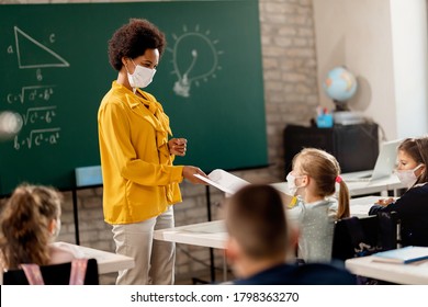 Happy black teacher and her students wearing protective face mask in the classroom. Teacher is giving them their test results. 