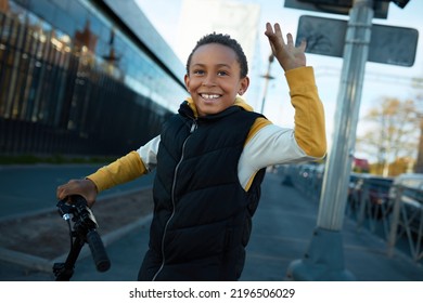 Happy black teen boy waving hand doing hi gesture greeting his friend, driving scooter on city streets, going to skate park to train new skills and tricks. Happy active childhood. Urban activity - Powered by Shutterstock