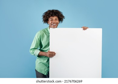 Happy Black Teen Guy Holding Empty Paper Poster With Mockup For Your Ad Design On Blue Studio Background. Excited African American Teenager Presenting Empty Banner