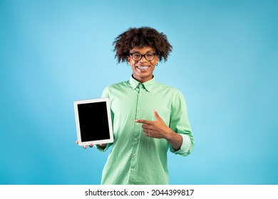 Happy Black Teenager Pointing At Tablet Computer With Mockup For Website Or App On Screen, Standing Over Blue Studio Background. Teen Guy Showing Touch Pad With Space For Your Ad