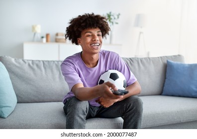 Happy Black Teenager Watching Football Game On TV At Home. Teen Sports Fan Sitting On Sofa In Living Room, Enjoying Television Game Match, Following His Favorite Soccer Team During Championship